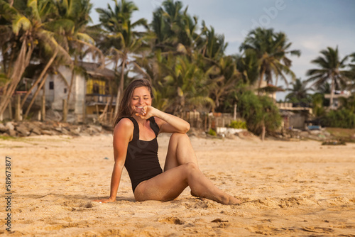 Happy smiling pretty woman in swimsuit on sandy beach at tropical palm trees background, summer vacation. Lady posing on tropical seacoast, relaxing lifestyle. Travel holiday concept. Copy text space