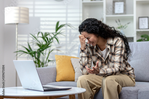 Online consultation with a psychologist, woman crying at home complaining to doctor on video call, Hispanic woman with curly hair talking to support service using laptop sitting on sofa at home.