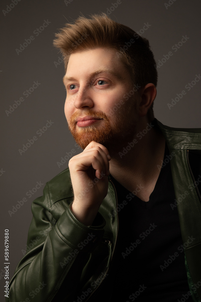 Studio portrait of an attractive caucasian man in his early 20s. He is wearing a green leather jacket. He has strawberry blonde hair and a beard. Studio portrait of an attractive caucasian man in his early 20s. He is wearing a green leather jacket. He has strawberry blonde hair and a beard.