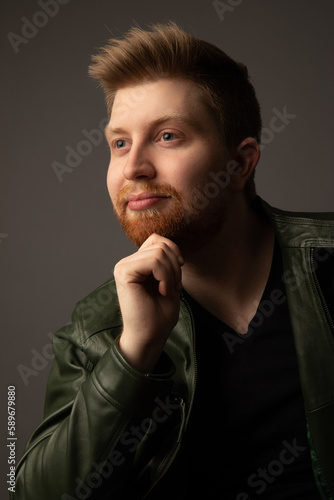 Studio portrait of an attractive caucasian man in his early 20s. He is wearing a green leather jacket. He has strawberry blonde hair and a beard. 