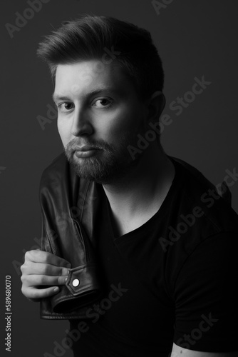 Studio portrait of an attractive caucasian man in his early 20s. He is wearing a leather jacket. He has strawberry blonde hair and a beard. This photo is black and white.