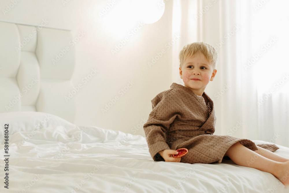 Portrait of a boy in a brown bathrobe, who is sitting on a white bed with candied oranges in his hands Portrait of a boy in a brown bathrobe, who is sitting on a white bed with candied oranges in his hands