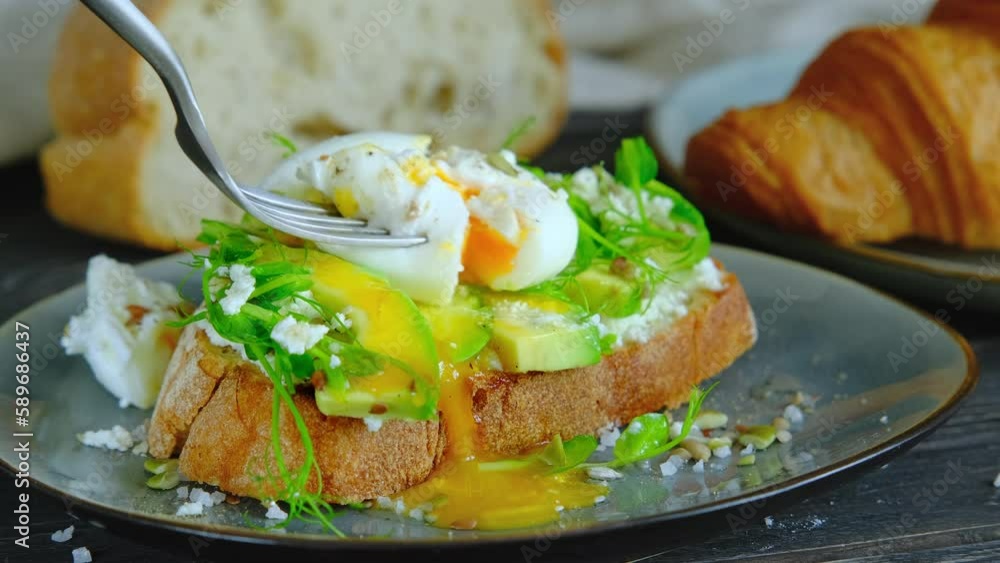 Person eating a poached egg on avocado toast, cutting a benedict egg with avocado spread on sandwich with fork, runny egg yolk, healthy breakfast, close-up video clip, 4k high quality footage Person eating a poached egg on avocado toast, cutting a benedict egg with avocado spread on sandwich with fork, runny egg yolk, healthy breakfast, close-up video clip, 4k high quality footage
