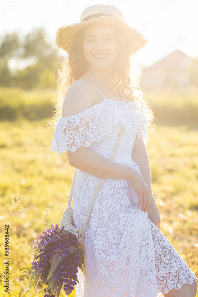 Beautiful young girl in a white dress, straw hat, with picnic and bouquet of purple wild flowers on a meadow. Summertime, golden hour, sunset. Wellness, wellbeing. Calendar
