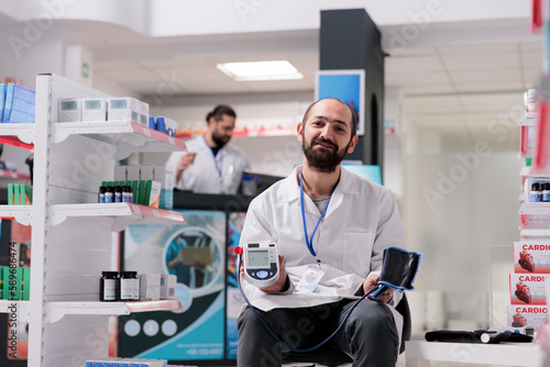 Portrait of pharmacist holding medical tonometer ready to measure clients hypertension during checkup visit in pharmacy. Drugstore filled with supplements and vitamins  medicine service