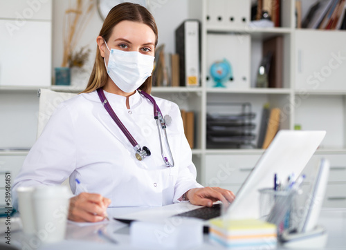 Portrait of young female doctor in face mask working on laptop in clinic office