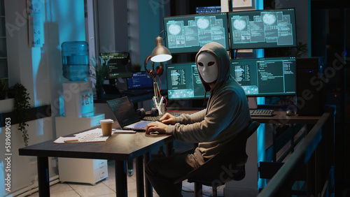 Male criminal wearing mask and hood to hack computer system, breaking into company servers to steal big data. Masked man looking dangerous and scary, impostor creating security malware. Handheld shot. photo