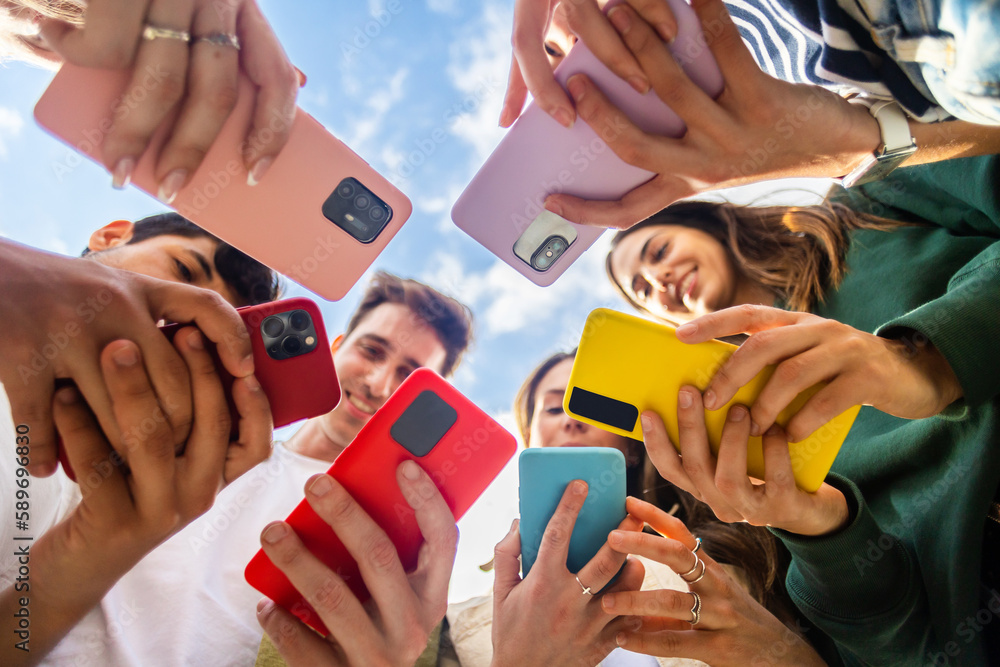 Young group of people using mobile phone device standing together in circle outdoors. Millennial friends addicted to social media app, betting or playing video game on platform online. - obrazy, fototapety, plakaty 