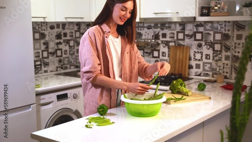 A Young smiling beautiful woman in oversize pink shirt in the kitchen table at home taken a salad spinner with helda beans and touches fresh green pods. Concept healthy nutrition and veggie lifestyle. photo