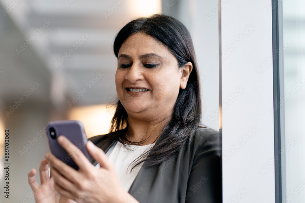 Smiling mature Indian woman holding smartphone using mobile app shopping online. Positive asian female communication, reading text message indoors. Mobile banking, technology concept Smiling mature Indian woman holding smartphone using mobile app shopping online. Positive asian female communication, reading text message indoors. Mobile banking, technology concept