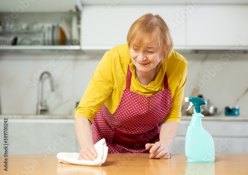 Mature woman in an apron, who cleaning the home kitchen with cleaning products, wipes the table with a damp cloth