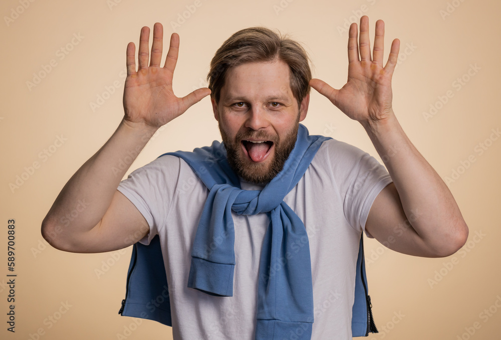 Comical funny young caucasian man making playful silly facial expressions and grimacing, fooling around, showing tongue. Handsome crazy adult guy bullying isolated on studio beige background indoors Comical funny young caucasian man making playful silly facial expressions and grimacing, fooling around, showing tongue. Handsome crazy adult guy bullying isolated on studio beige background indoors