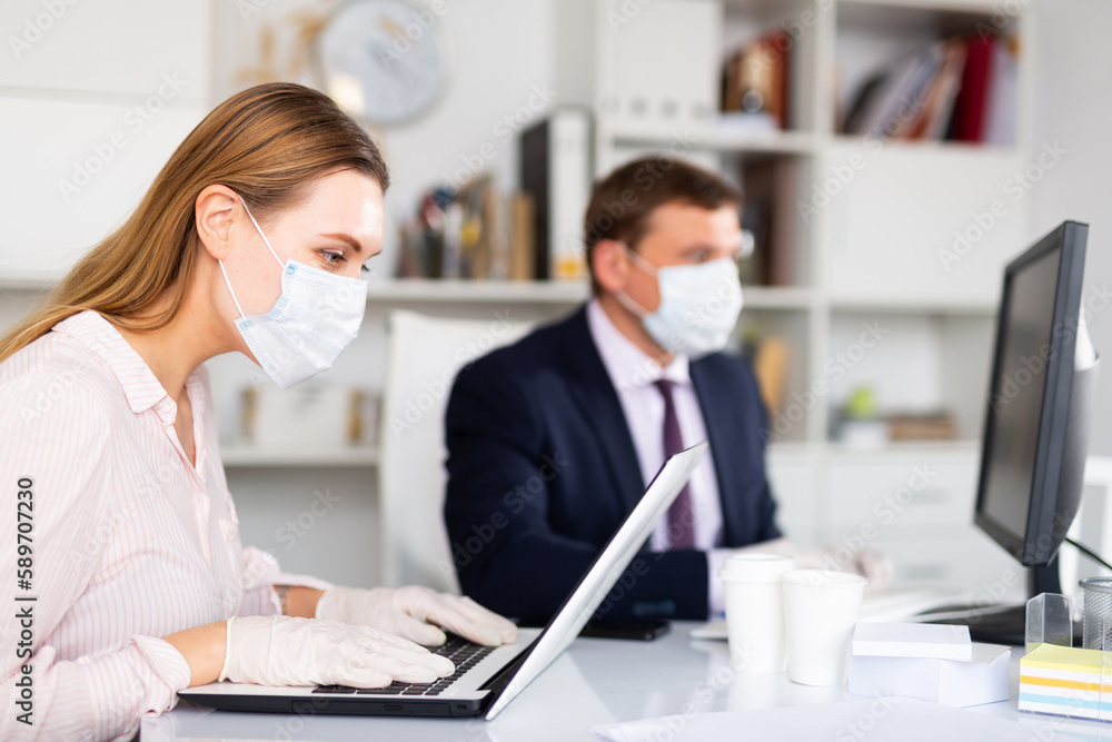 Obraz premium Focused young woman in medical face mask and latex gloves working on laptop in office with male colleague. Concept of prevention of viral infection and social distancing