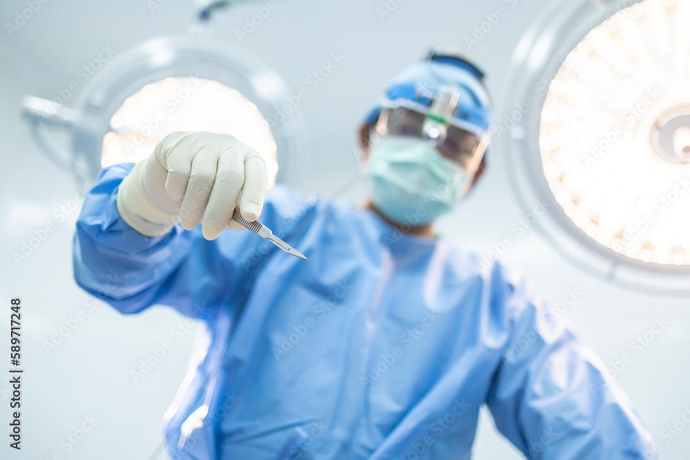 Uprisen of surgeon or doctor holding scalpel inside operating room in hospital with blurred surgical lamp background.Surgeon in blue uniform did surgery technology.Selective focus at hand of people. - obrazy, fototapety, plakaty 