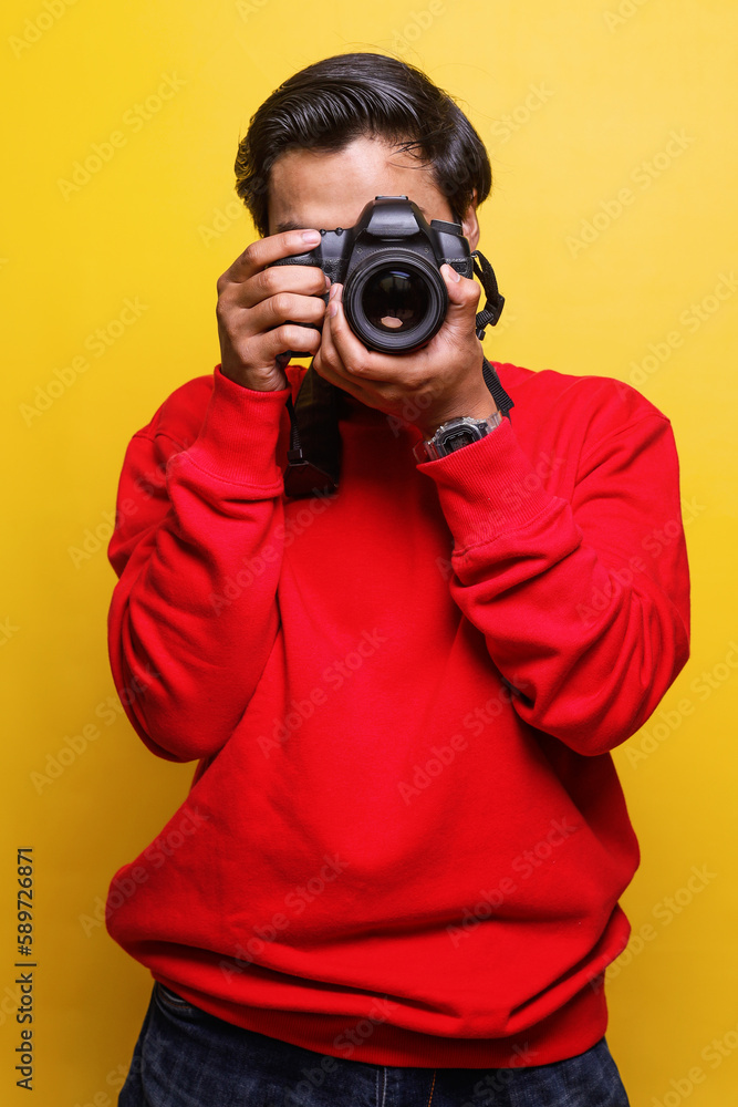 Young professional photographer in red shirt take picture of you using camera isolated on yellow color background Young professional photographer in red shirt take picture of you using camera isolated on yellow color background