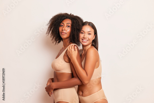 Two lovely model girls in beige underwear are standing embracing on white background, happy African American and Asian young woman, friendship concept, copy space, high quality photo