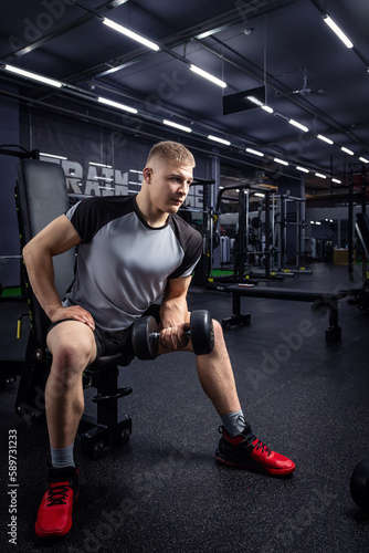 A athlete with blond hair shakes biceps in dumbbells in the gym.