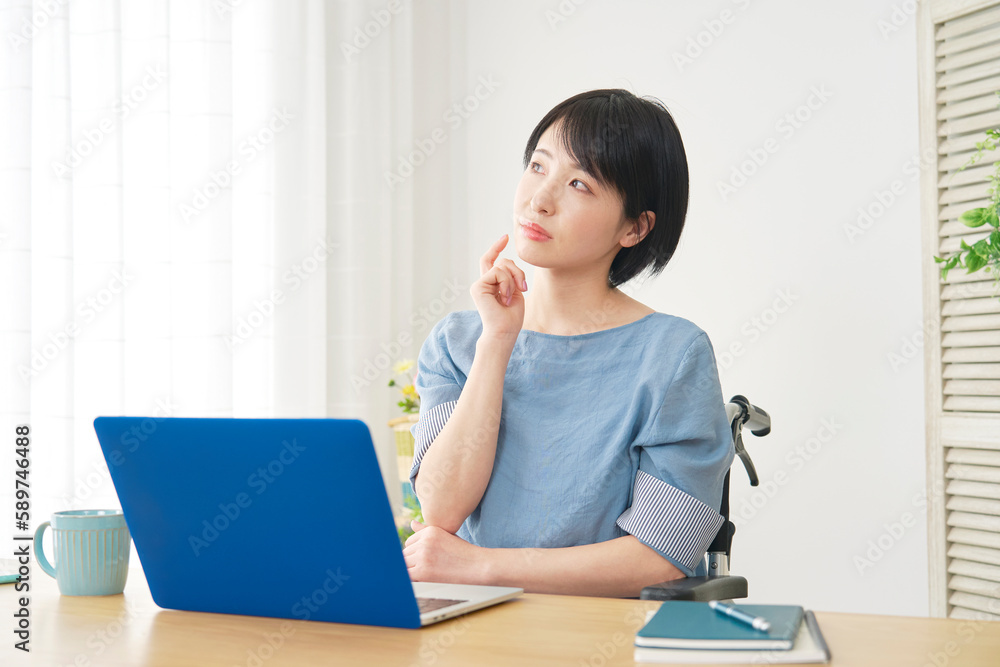 リビングでノートパソコンを使いながら考える車椅子に乗った女性 リビングでノートパソコンを使いながら考える車椅子に乗った女性