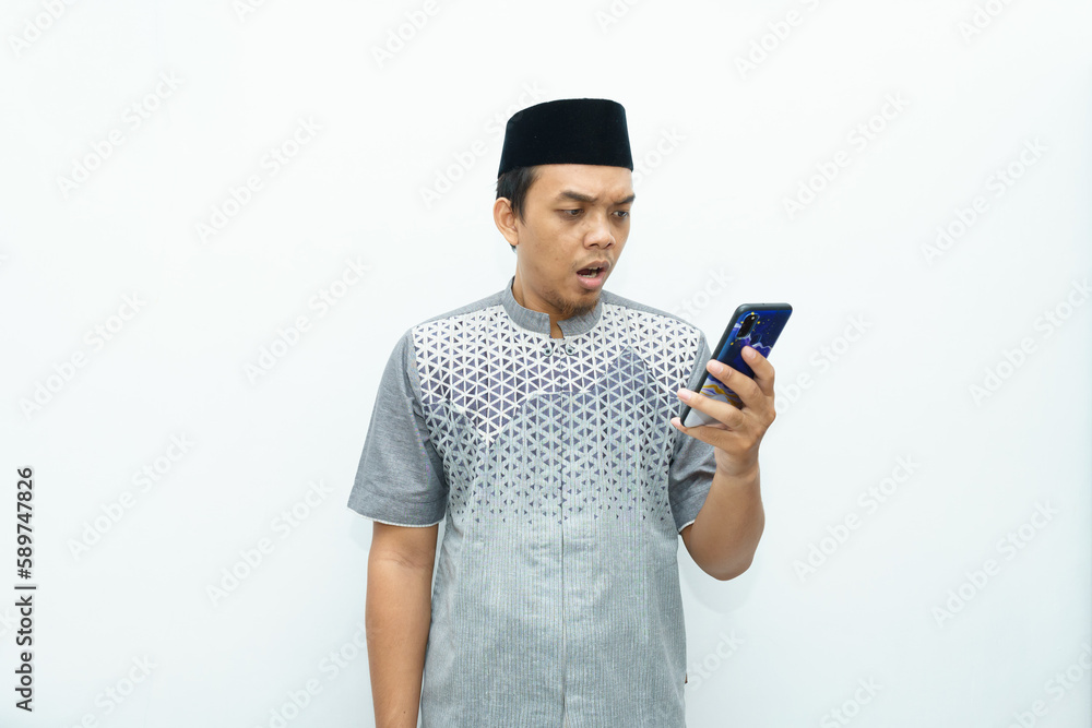 Portrait Indonesian Asian muslim man shocked looking at smartphone wears black hat called peci or songkok Portrait Indonesian Asian muslim man shocked looking at smartphone wears black hat called peci or songkok