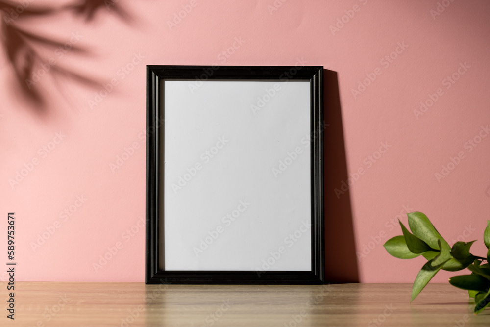 Black empty frame with copy space with plant on table against pink wall and shadow of leaves Black empty frame with copy space with plant on table against pink wall and shadow of leaves