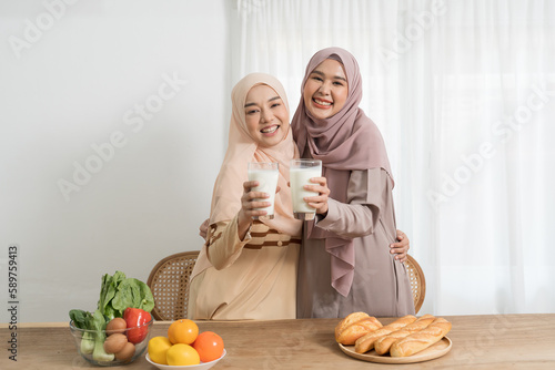 Canvas Print Two young Asian muslim women wear hijab drinking a glass of milk with happy and smiling in kitchen at home
