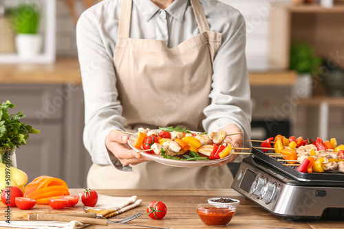 Woman holding plate with grilled chicken skewers and vegetables in kitchen  closeup