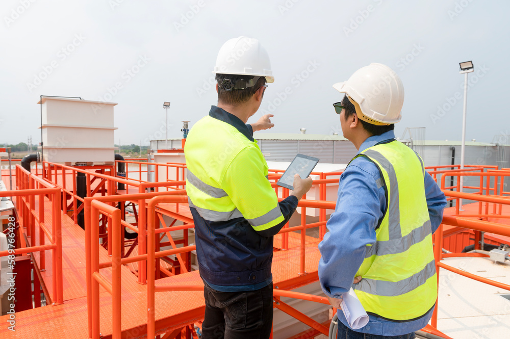 Two heavy industrial engineers standing in a standard water treatment plant use digital tablet computers and chatting. Asian industry professionals Two heavy industrial engineers standing in a standard water treatment plant use digital tablet computers and chatting. Asian industry professionals