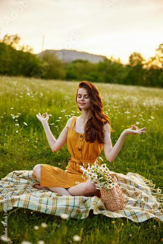 a red-haired woman in an orange dress sits on a plaid in a chamomile field and midit in the lotus position