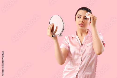 Young woman with mirror removing makeup on pink background