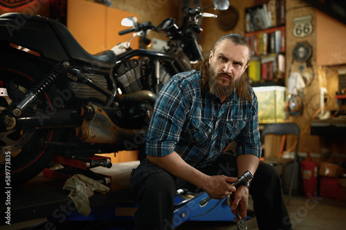 Portrait of mature bearded biker looking at camera sitting in his own garage