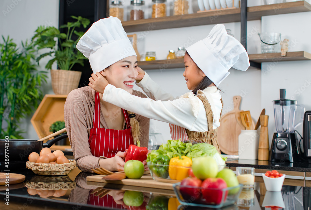 Millennial Asian young little cute girl chef daughter in apron standing smiling wearing adjusting white tall cook hat for mother ready for cooking vegetables food meal at counter in home kitchen Millennial Asian young little cute girl chef daughter in apron standing smiling wearing adjusting white tall cook hat for mother ready for cooking vegetables food meal at counter in home kitchen