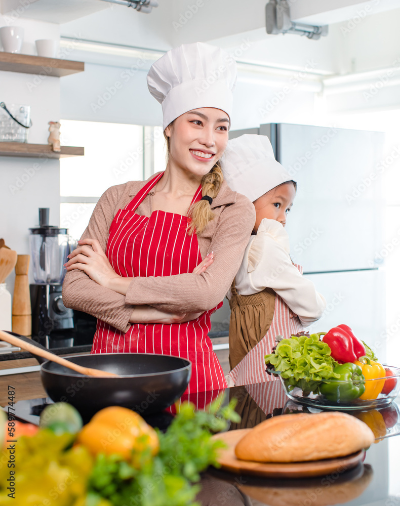 Asian young cheerful female mother and little cute girl daughter wearing white tall cook hat and apron standing crossed arms smiling posing holding thumbs up together ready for cooking in kitchen Asian young cheerful female mother and little cute girl daughter wearing white tall cook hat and apron standing crossed arms smiling posing holding thumbs up together ready for cooking in kitchen