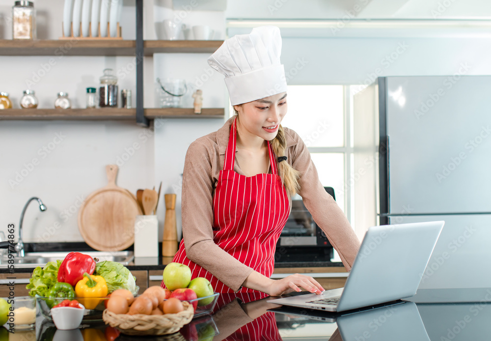 Portrait shot of Asian young cute female chef wears white tall cook hat and apron smiling posing look at camera learning cooking homemade food meal online via laptop notebook computer in home kitchen Portrait shot of Asian young cute female chef wears white tall cook hat and apron smiling posing look at camera learning cooking homemade food meal online via laptop notebook computer in home kitchen