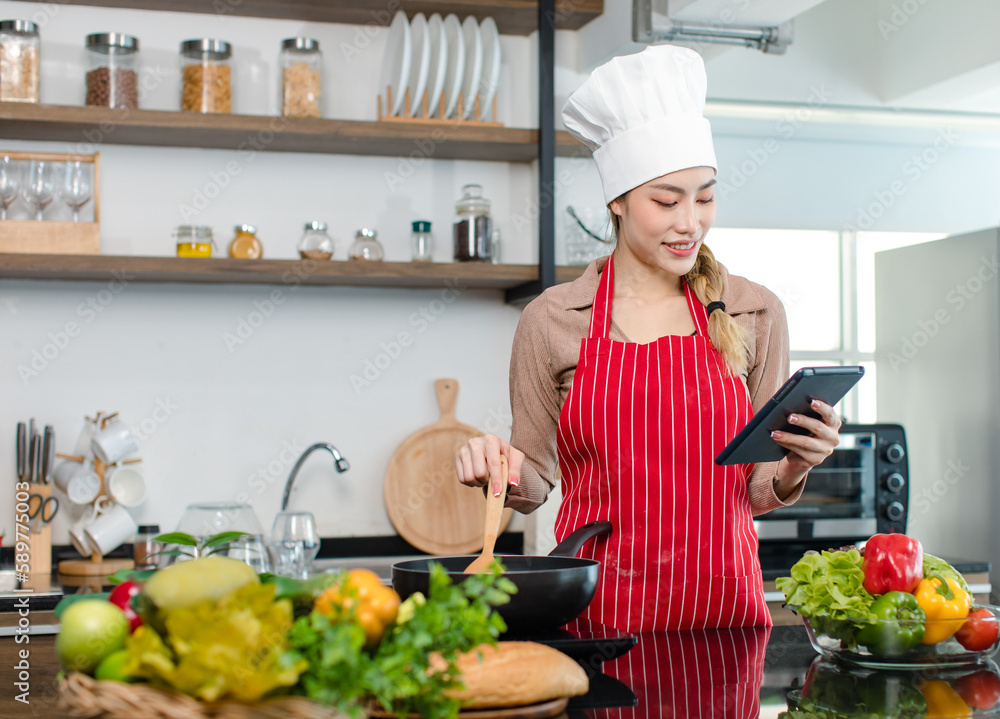 Asian young female chef housewife wears white tall cook hat and apron learning cooking homemade food meal online via touchscreen tablet computer while using spatula frying eggs in pan at home kitchen Asian young female chef housewife wears white tall cook hat and apron learning cooking homemade food meal online via touchscreen tablet computer while using spatula frying eggs in pan at home kitchen