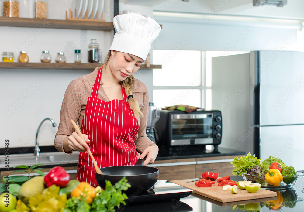 Asian young female chef housewife wears white tall cook hat and apron standing holding wooden spatula spoon tasting food meal from frying pan while cooking at counter with vegetables in home kitchen Asian young female chef housewife wears white tall cook hat and apron standing holding wooden spatula spoon tasting food meal from frying pan while cooking at counter with vegetables in home kitchen
