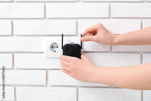 Woman plugging black WiFi repeater in electric socket on white brick wall