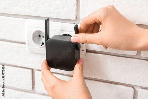 Woman plugging black WiFi repeater in electric socket on white brick wall