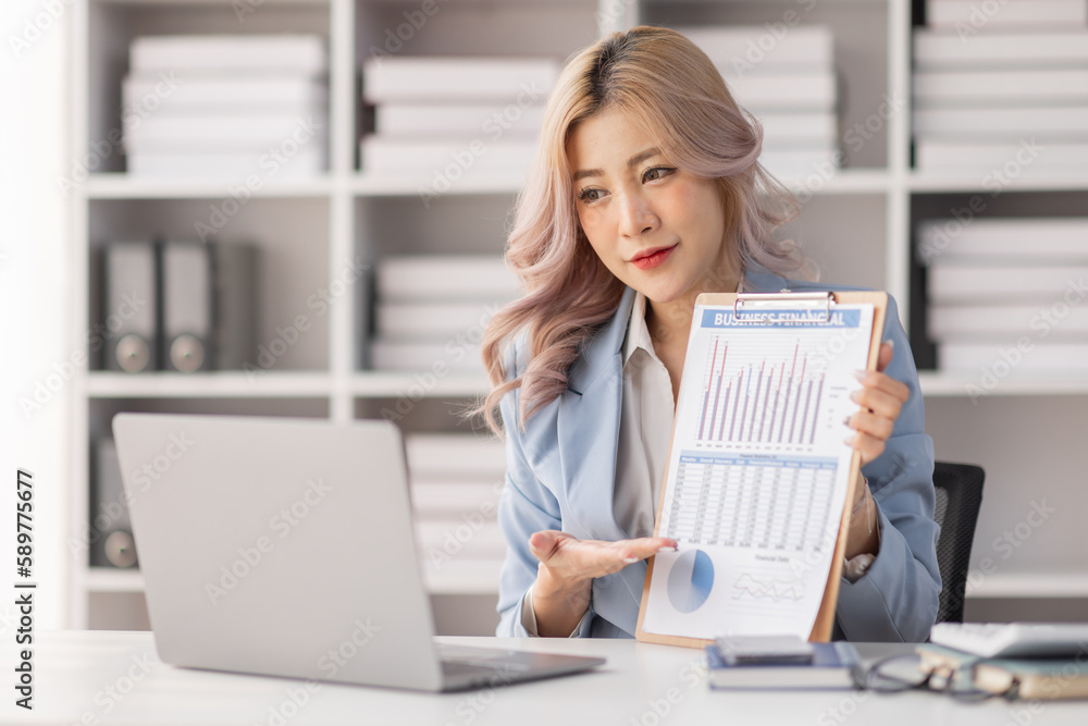 A young Asian businesswoman shows documents with graphs with a pen, holds in his hand smiles. Sitting at a desk with a laptop in a modern office. A young Asian businesswoman shows documents with graphs with a pen, holds in his hand smiles. Sitting at a desk with a laptop in a modern office.