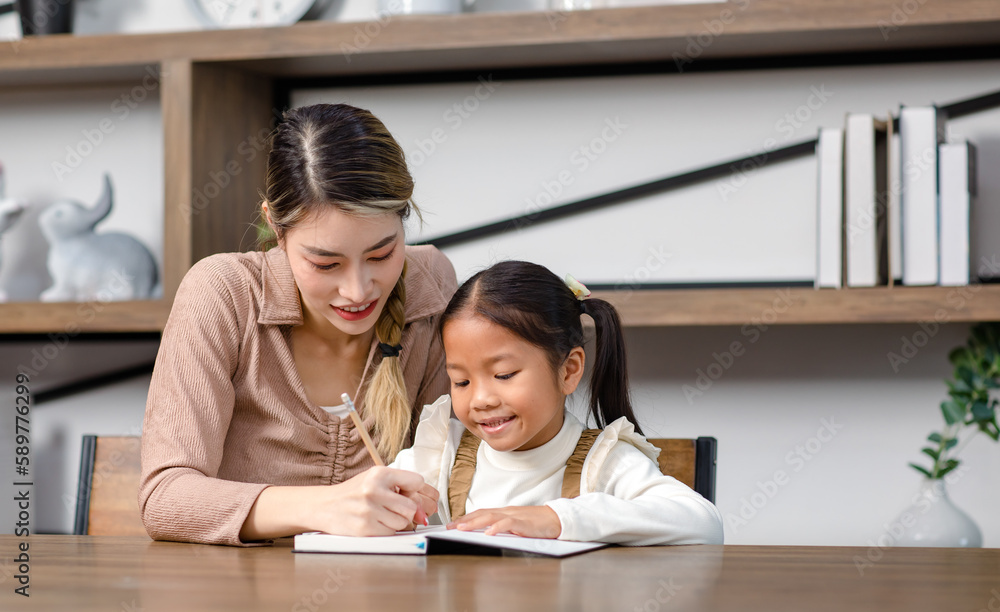 Asian young female housewife mother tutor teacher sitting smiling on table in living room at home teaching little cute kindergarten preschool girl daughter writing on book doing homework after school Asian young female housewife mother tutor teacher sitting smiling on table in living room at home teaching little cute kindergarten preschool girl daughter writing on book doing homework after school