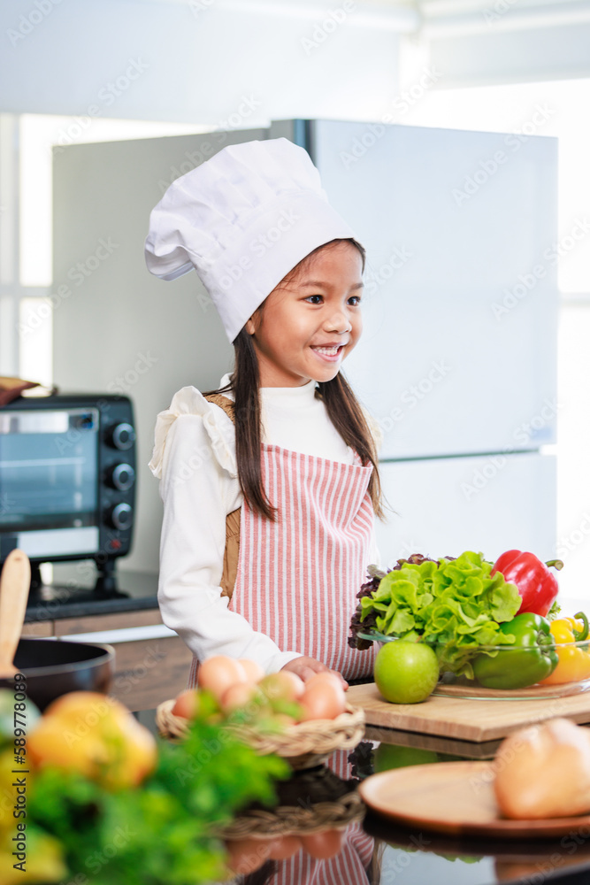 Millennial Asian young little cute girl chef daughter with white tall cook hat and apron standing smiling holding wooden spoon and fork posing ready for cooking vegetables at counter in home kitchen Millennial Asian young little cute girl chef daughter with white tall cook hat and apron standing smiling holding wooden spoon and fork posing ready for cooking vegetables at counter in home kitchen