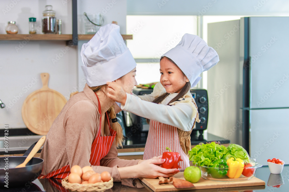 Millennial Asian young little cute girl chef daughter in apron standing smiling wearing adjusting white tall cook hat for mother ready for cooking vegetables food meal at counter in home kitchen Millennial Asian young little cute girl chef daughter in apron standing smiling wearing adjusting white tall cook hat for mother ready for cooking vegetables food meal at counter in home kitchen