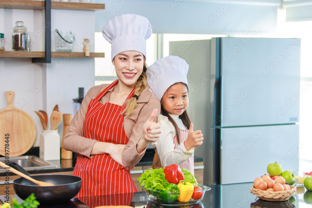 Asian young cheerful female mother and little cute girl daughter wearing white tall cook hat and apron standing crossed arms smiling posing holding thumbs up together ready for cooking in kitchen Asian young cheerful female mother and little cute girl daughter wearing white tall cook hat and apron standing crossed arms smiling posing holding thumbs up together ready for cooking in kitchen