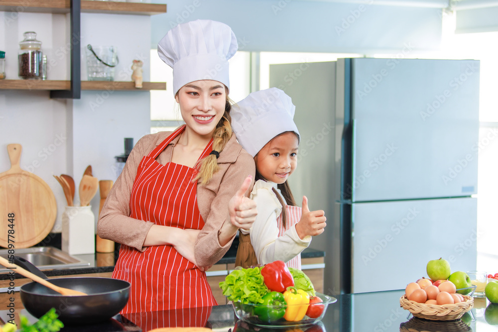 Asian young cheerful female mother and little cute girl daughter wearing white tall cook hat and apron standing crossed arms smiling posing holding thumbs up together ready for cooking in kitchen Asian young cheerful female mother and little cute girl daughter wearing white tall cook hat and apron standing crossed arms smiling posing holding thumbs up together ready for cooking in kitchen