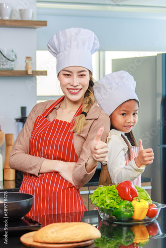 Asian young cheerful female mother and little cute girl daughter wearing white tall cook hat and apron standing crossed arms smiling posing holding thumbs up together ready for cooking in kitchen