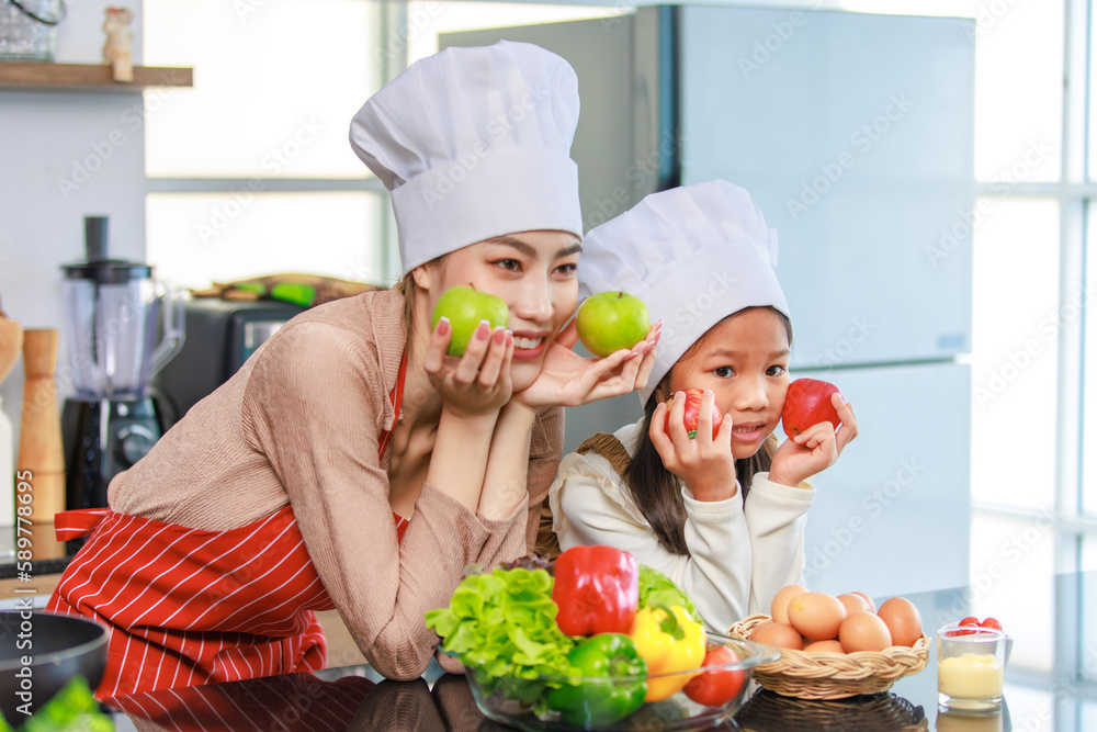 Asian young female chef housewife mother and little cute girl daughter wears white tall cook hat and apron smiling posing looking at home kitchen. Asian young female chef housewife mother and little cute girl daughter wears white tall cook hat and apron smiling posing looking at home kitchen.