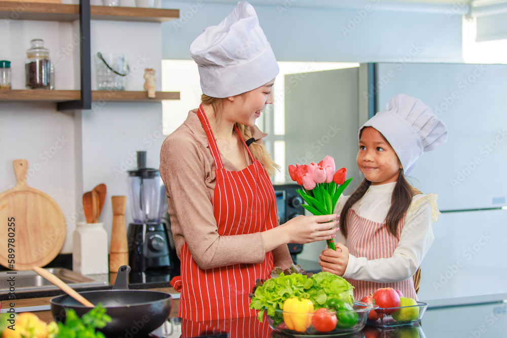 Asian young cheerful female chef housewife mother and little girl daughter wears white tall cook hat and apron standing smiling holding tulip flower bouquet together in kitchen preparing food meal Asian young cheerful female chef housewife mother and little girl daughter wears white tall cook hat and apron standing smiling holding tulip flower bouquet together in kitchen preparing food meal