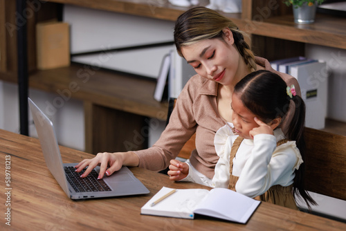Asian young female housewife mother tutor teacher sitting smiling on table in living room using notebook computer pointing teaching little cute kindergarten preschool girl daughter doing homework
