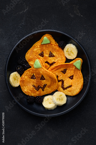 Jack-o'-lantern pumpkin pancakes with chocolate hazelnut spread and banana on a black background. Dessert for halloween. Top view