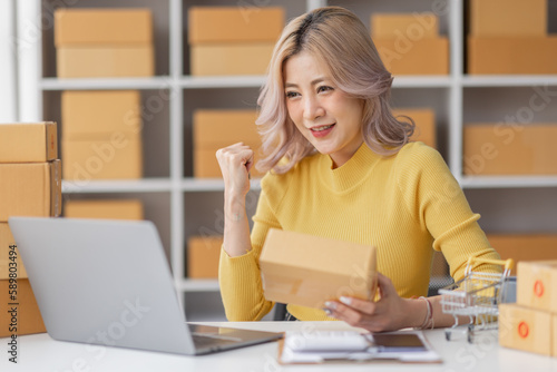 Woman in a small start-up business woman working in a home office working on a laptop and smartphone checking orders from the internet in preparation for shipping. 