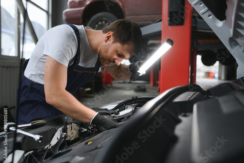 Handsome young male auto mechanic in special uniform clothes holding a flashlight, looking for breakdown and repairing under the hood in the car engine in a car workshop